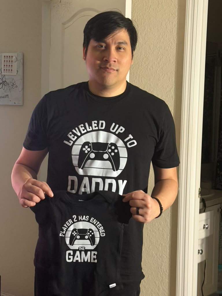 Mikey holding a baby onesie next to his Leveled Up To Daddy shirt
